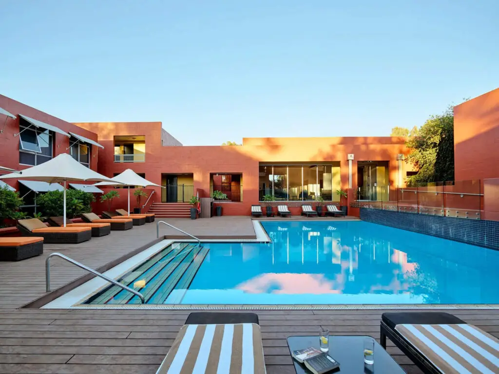 An outdoor pool area at dusk within a hotel complex, showing the reflective blue water surrounded by loungers and umbrellas. The surrounding buildings have a warm, terracotta colour, and trees line the backdrop under a fading blue sky. The Lost Camel is one of the more affordable Uluru accommodation options - and has a bit of personality and colour to match!