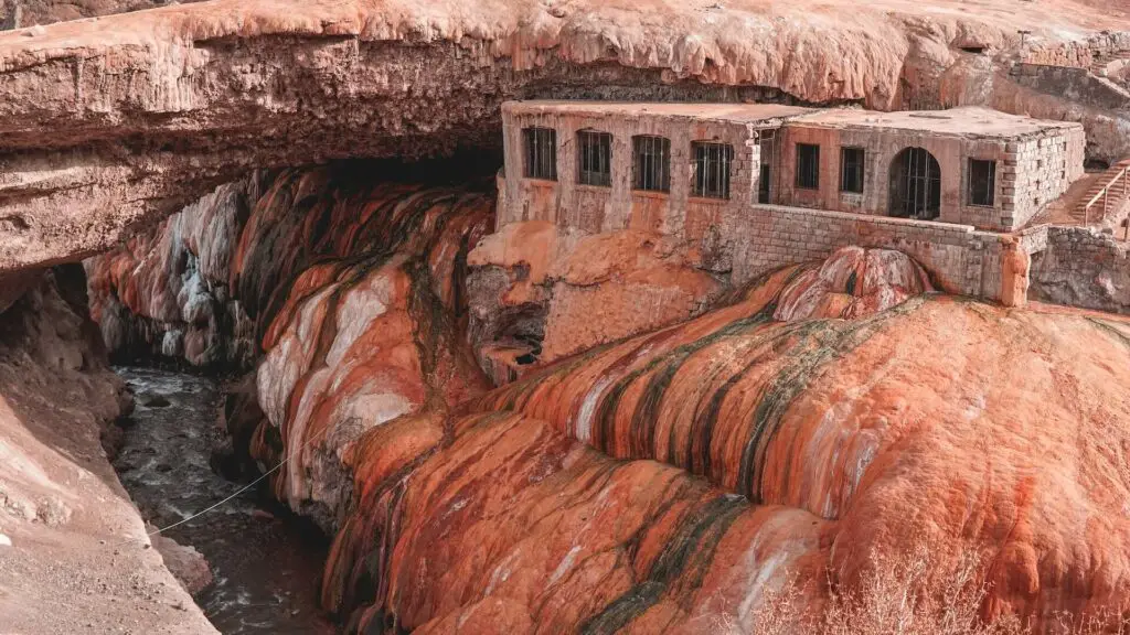 An abandoned brick or stone building perched over a river of red-tinged water flowing through a rugged terrain. This is the Puente del Inca in Mendoza, Argentina.