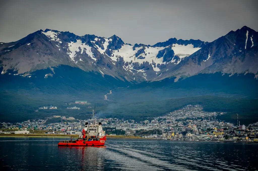 A red tug boat glides on the water in front of Ushuaia, a great destination for honeymooners visiting Argentina. Behind the town are snow-capped mountains.