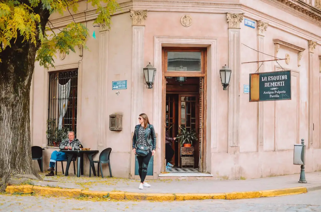 A quaint street corner with an old-fashioned building featuring a sign that reads "La Esquina de Merti." A woman walks by, and a man sits at a cafe table outside under the yellowing leaves of a tree.