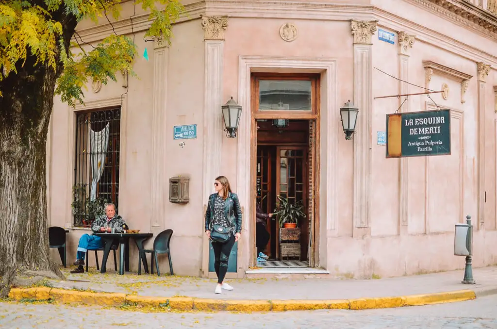 A quaint corner building in the historic town of San Antonio de Areco in Argentina, painted in light pink with ornate architectural details. A sign reads "La Esquina de Merti." A woman - the author of this article - walks past the open doorway, while a man sits at an outdoor table beneath a large tree.