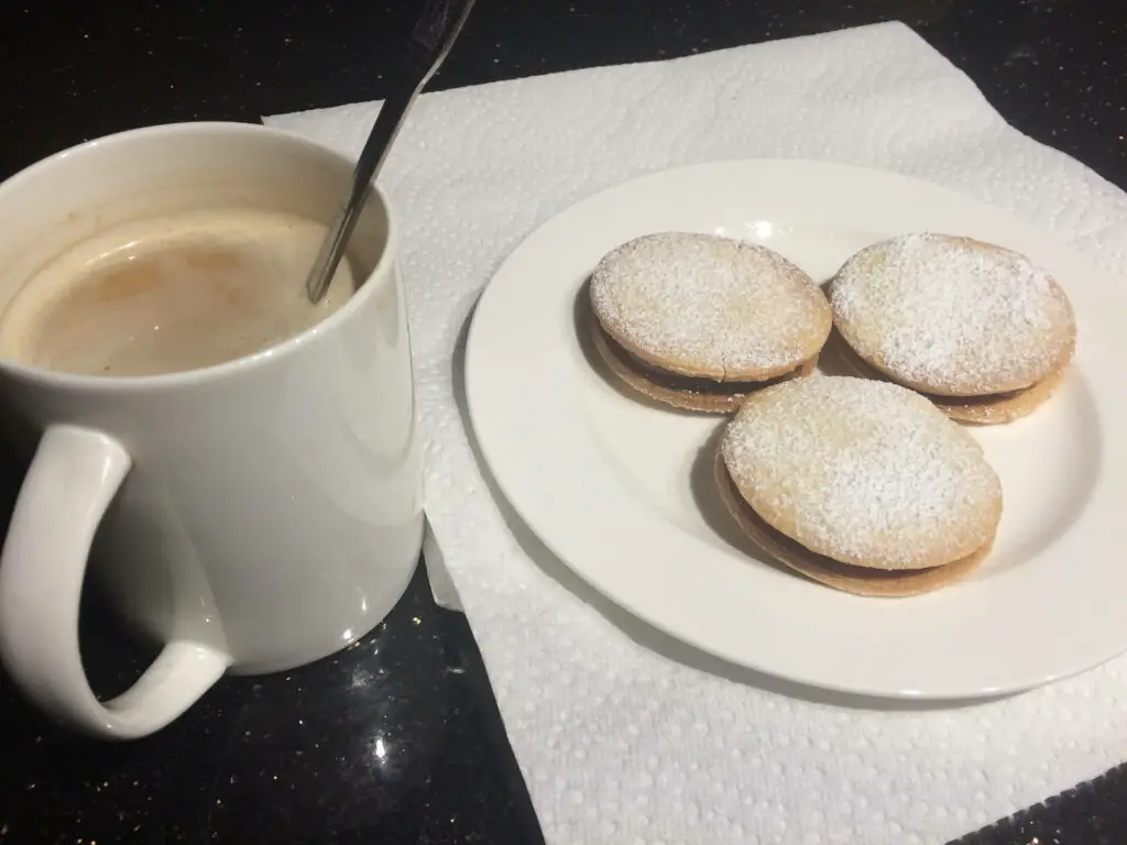 A white mug of coffee with a spoon inside is on the left, and a plate with three powdered sugar-coated cookies called alfajores is on the right, both on a dark countertop. Alfajores are one of the traditional foods in Argentina.