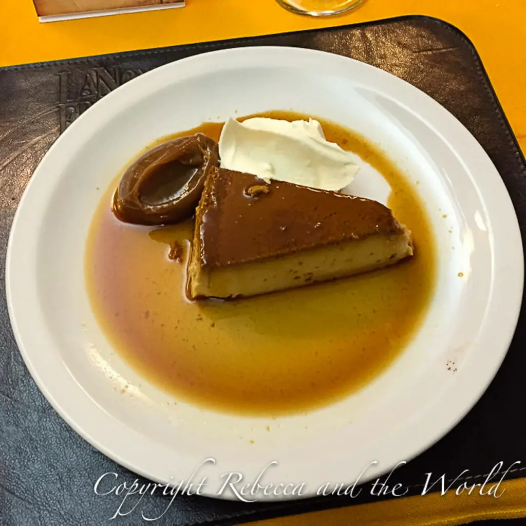 A caramel flan dessert on a white plate garnished with a dollop of whipped cream and a dollop of dulce de leche, placed on a table with a brown leather mat.