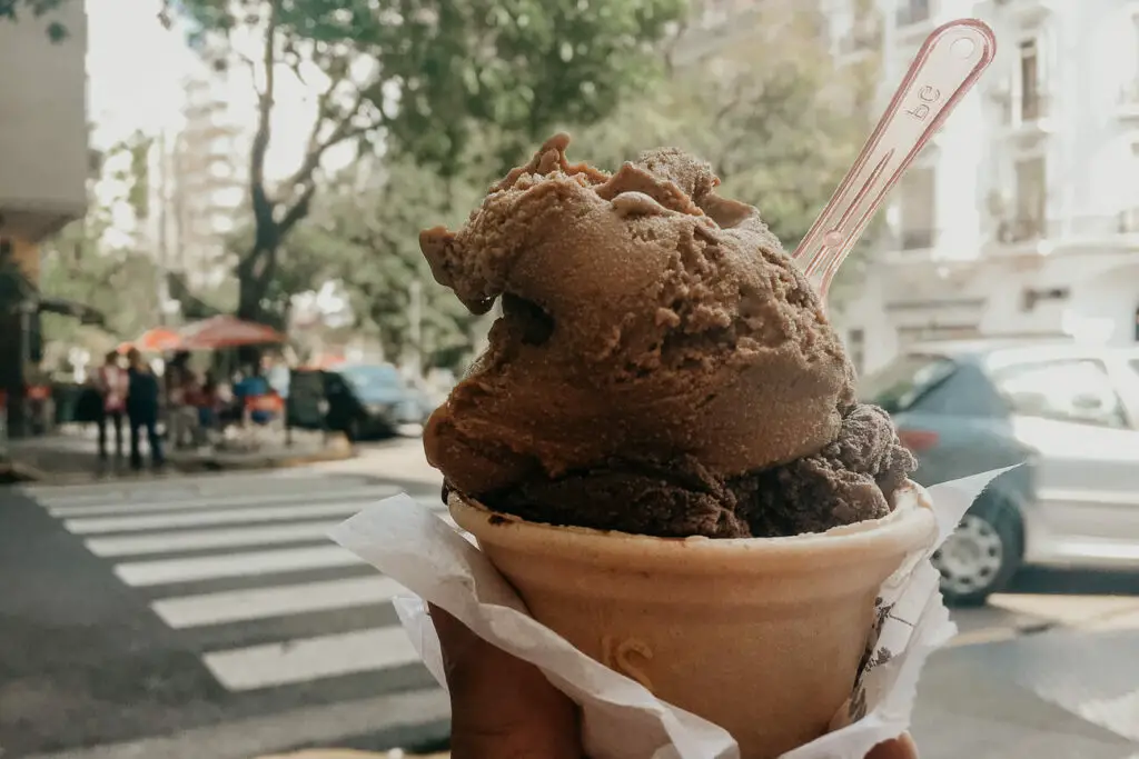 A hand holding a paper cup filled with chocolate ice cream, with a clear plastic spoon in it. The background shows a city street scene in soft focus. Ice cream is one of the best foods in Argentina.