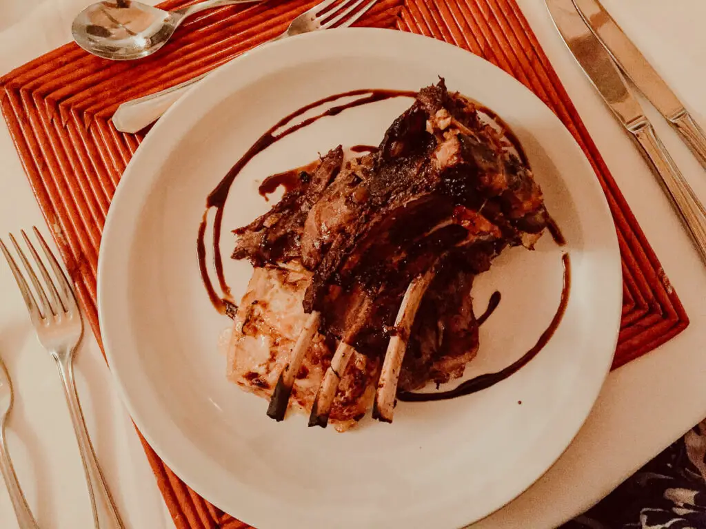 A roasted rack of lamb with herbs and a balsamic reduction drizzle on a white plate, against a background with red stripes, suggesting a restaurant setting. Patagonian lamb is a typical Argentinian food.