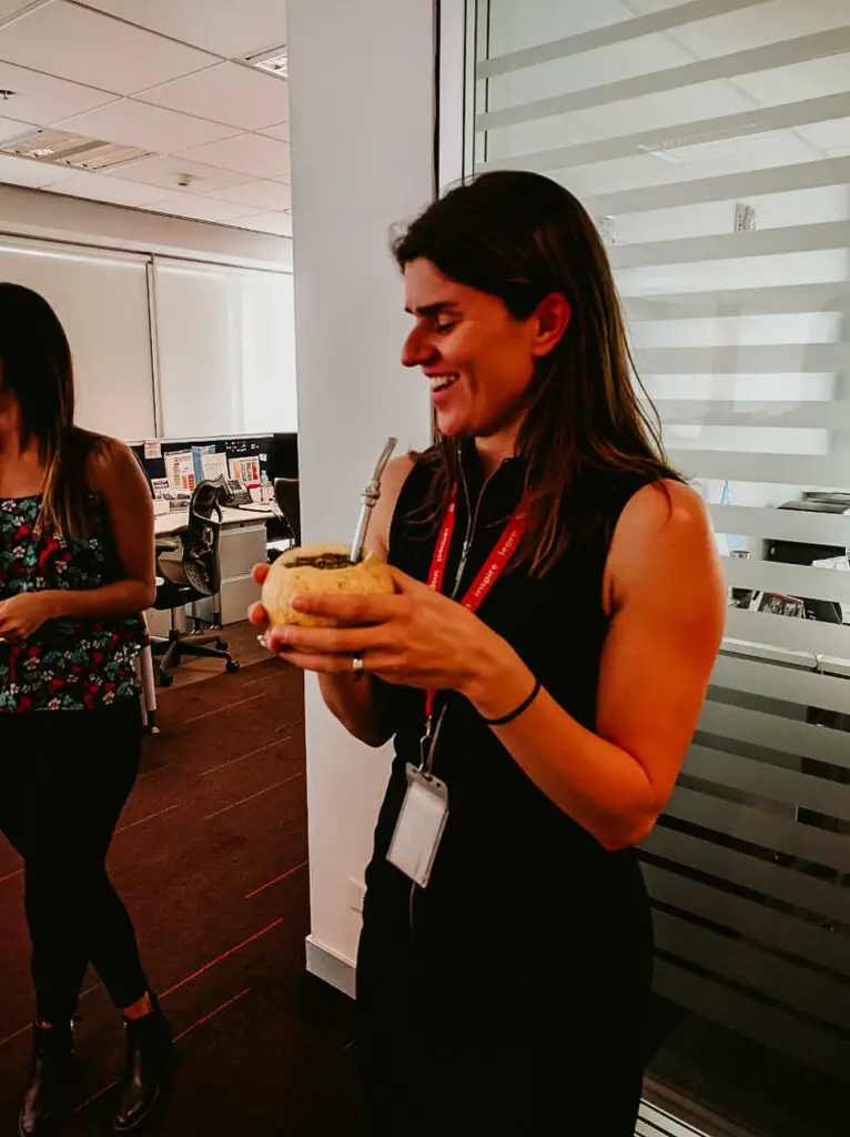 A smiling woman (the author of this article) in an office environment is holding a hollowed-out round grapefruit filled with mate with a bombilla (straw) sitting in the fruit.