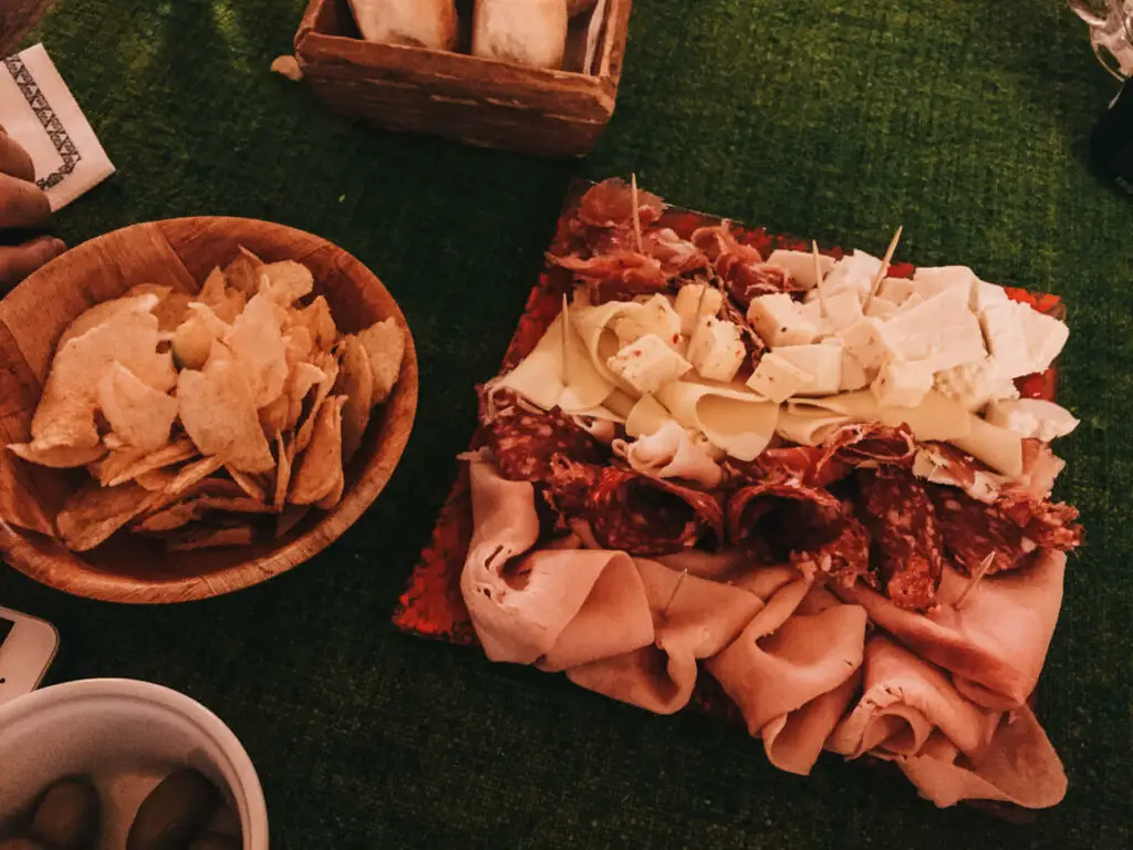 A picada (charcuterie) board with an assortment of sliced meats, cheeses, and a bowl of chips, presented on a table with a green covering, suggesting an informal gathering. Meals in Argentina usually start with picada.