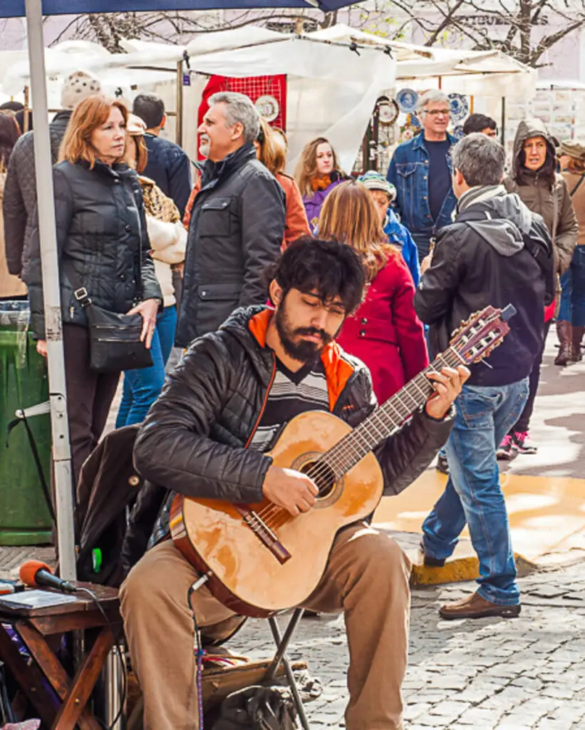 A man sits in a busy square in San Telmo, Buenos Aires. His eyes are closed and he is strumming a guitar. The passion and culture are reasons to visit Argentina.