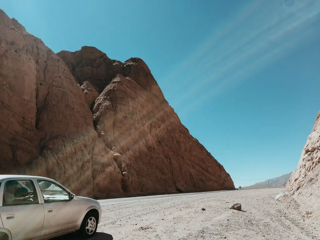 A grey car is parked on the side of the road with a rocky cliff looming over it.