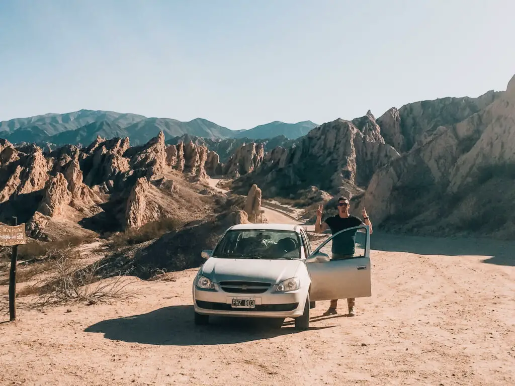 A man stands by a grey rental car with his fingers in the peace sign. Behind him are the craggy peaks of the Quebrada las Flechas in North Argentina. 