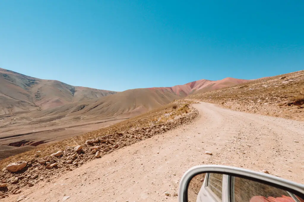 A dirt road in Argentina with hills in the background. In the foreground is the side mirror of a rental car