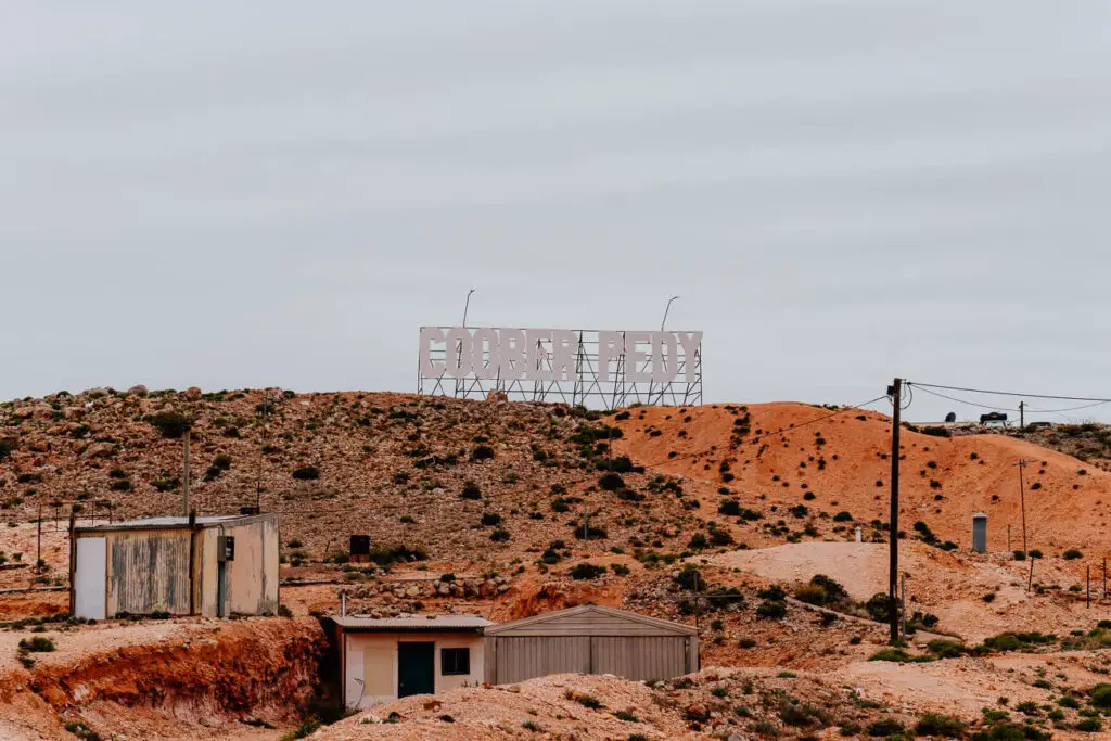 The Coober Pedy sign - in the design of the Hollywood sign - sits on top of an orange-red coloured hill. Shack-style buildings are in the foreground