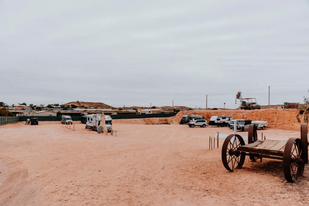 A barren and dusty open area has a handful of cars, caravans and motorhomes parked around in, including an old unused wagon in the foreground. This is the free camping site in Coober Pedy.