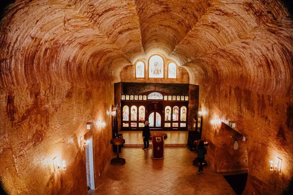 The inside of the Serbian Orthodox Church in Coober Pedy shows an ornately carved room built into a hill. A woman admires the leadlight features in the front of the church hall