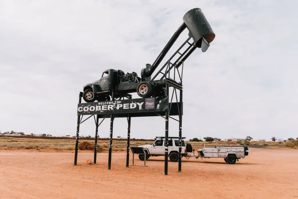 A white landcruiser towing a camper trailer is parked by the Welcome to Coober Pedy sign - a must-do in Coober Pedy. The sign is black with a truck on top
