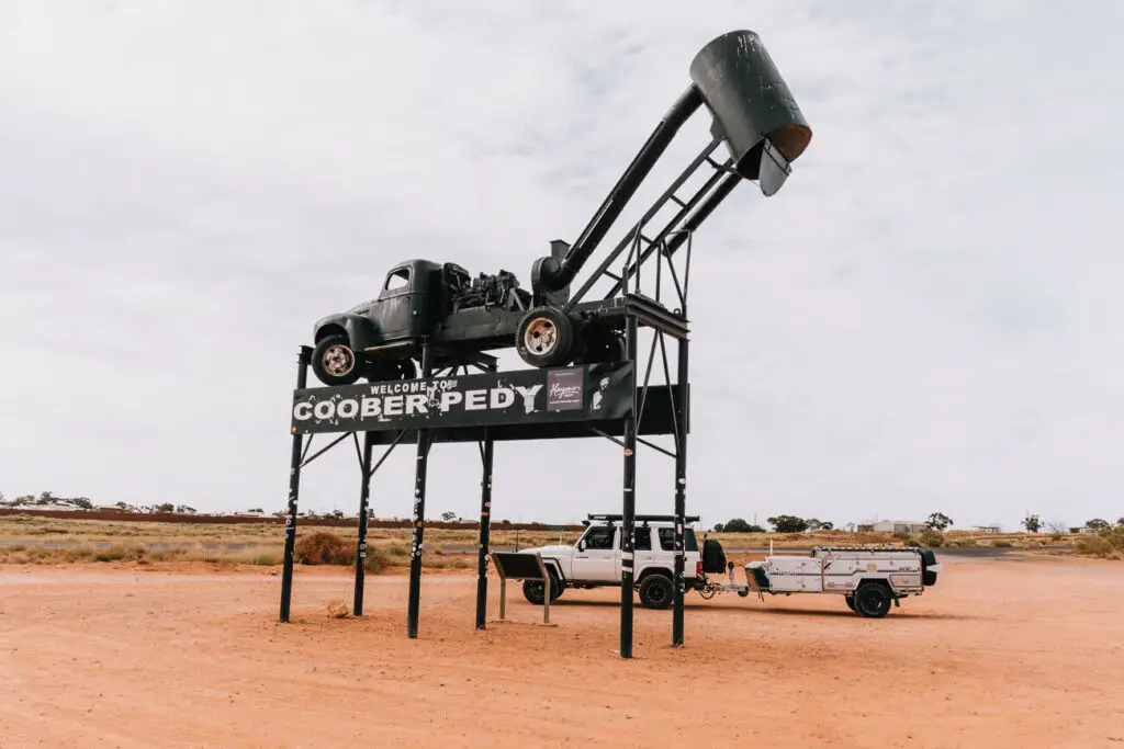 A classic car mounted high on a mining tower structure as a quirky welcome sign to the town of Coober Pedy, with flat arid land stretching into the distance.