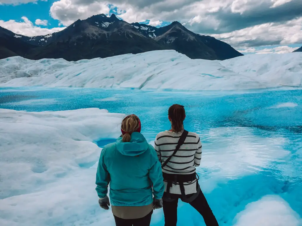 Two women with their backs to the camera look over a bright blue body of water on top of Perito Moreno Glacier in Argentina. Travel insurance for Argentina is important when tackling risky adventure activities like ice trekking.