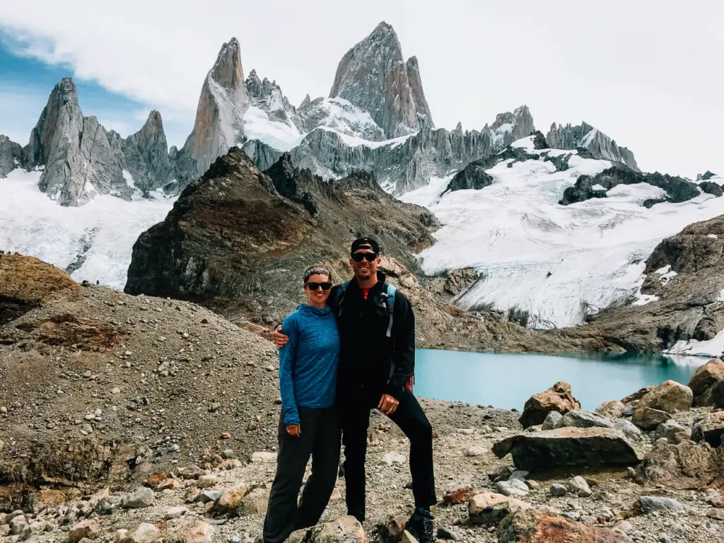 A man and woman (the author of this article) in hiking clothing smile at the camera as they stand in front of jagged, rocky, snow-capped mountain peaks in El Chalten, Argentina. Hiking and adventure activities are popular in Argentina so you should always buy travel insurance!