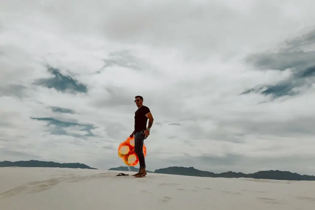 A man holding an orange sandboard stands on top of a sand dune at White Sands National Park in New Mexico