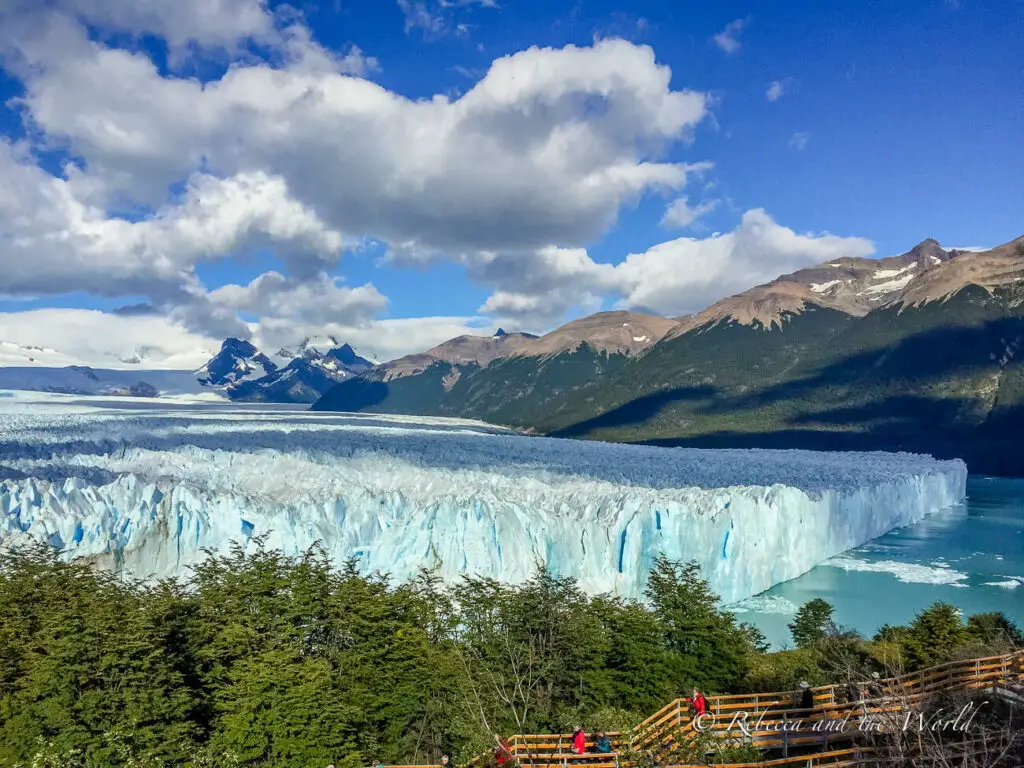 A vast glacier front with deep blue ice extending into a lake, flanked by forested hills and mountains under a partly cloudy sky, with tourists on a viewing platform to the left. This is Perito Moreno Glacier in Argentina.
