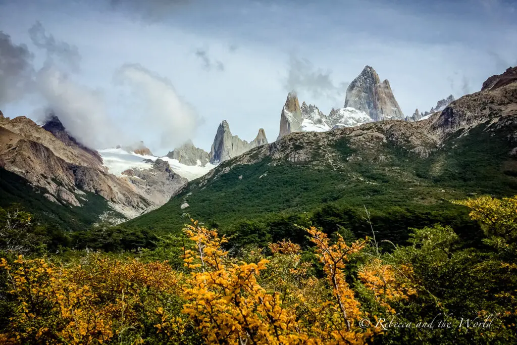 A majestic mountain range with sharp peaks, partially covered in snow, towering over a valley with orange and green foliage under a cloudy sky. This is Mount Fitz Roy in El Chalten, Argentina.