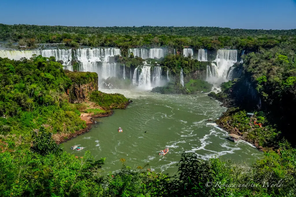 A panoramic view of the Iguazu Falls waterfall system with multiple cascades surrounded by lush greenery. Boats with tourists are visible on the river below. The waterfalls straddle the border of Argentina and Brazil in South America.