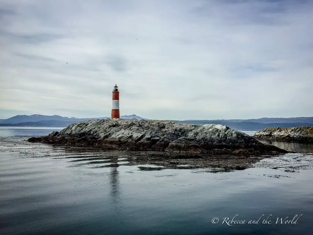 A solitary red and white lighthouse in Ushuaia, Argentina, on a small rocky outcrop, surrounded by calm seas with distant mountains under an overcast sky.