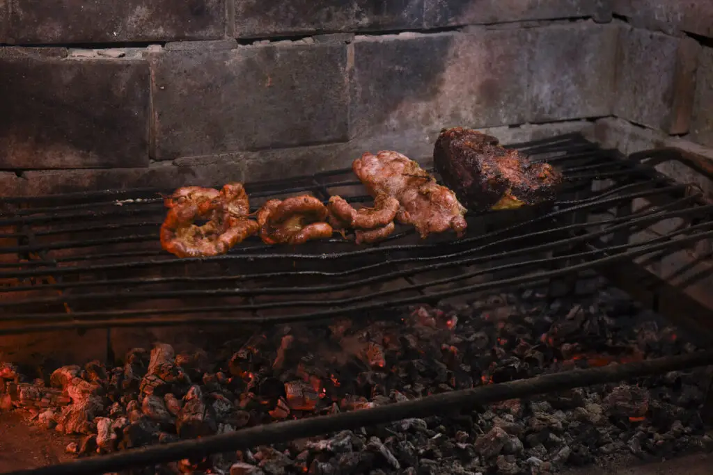 Chinchulines grilling on a barbecue in Argentina. The focus is on several pieces of meat grilling over glowing embers inside a brick barbecue pit. There are charred grill marks visible on the meat.