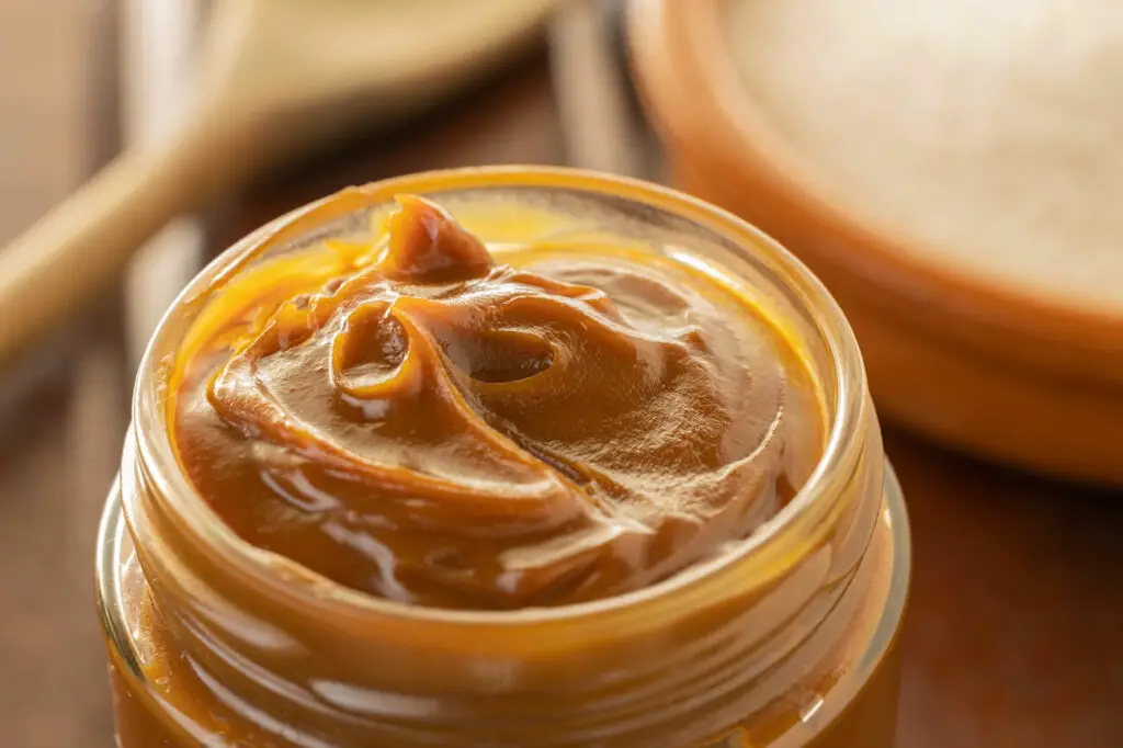 Close-up of a jar of Argentinian dulce de leche - a caramel sauce. A glass jar filled with thick, glossy caramel spread is in focus, with a wooden spoon and a container blurred in the background.