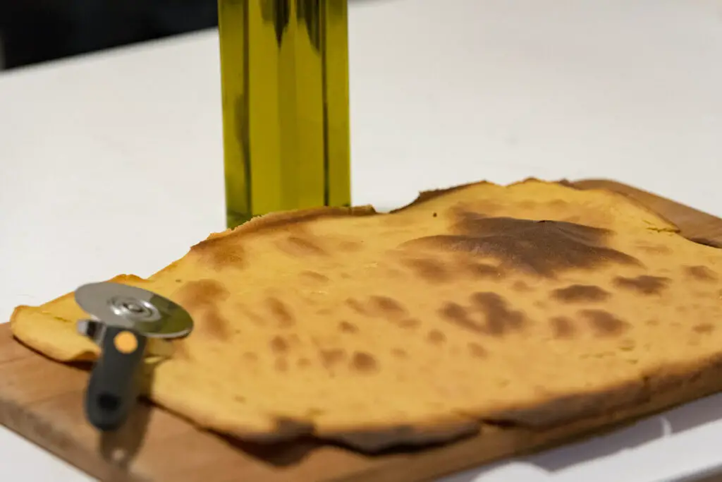 Faina - chickpea pizza - with a pizza cutter. The image shows a golden-brown flatbread with charred spots on a wooden board next to a pizza cutter and a bottle of olive oil, set against a white background.