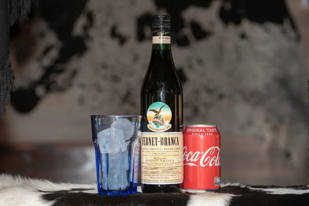 Beverages and ice in a glass. A bottle of Fernet-Branca, a can of Coca-Cola, and a blue glass filled with ice cubes are prominently displayed on a surface covered with a cow hide.