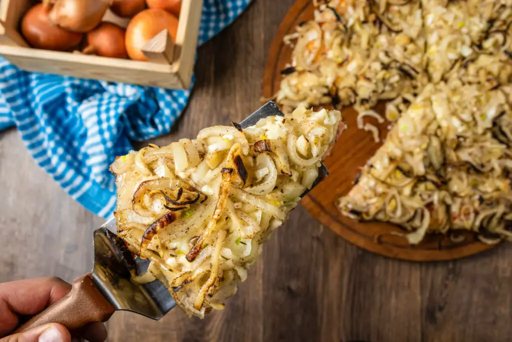 Pizza with onions, called fugazza in Argentina. A slice of pizza heavily topped with caramelized onions is being lifted from a wooden cutting board, with a basket of onions and a blue cloth in the background.