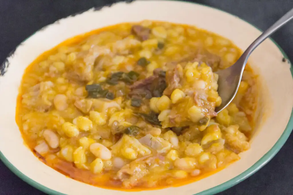 Locro in a bowl. A bowl of thick stew with white beans, corn, and leafy greens is visible, accompanied by a spoon, set on a striped black and white cloth. Locro is a traditional Argentinian dish that's filling and comforting.
