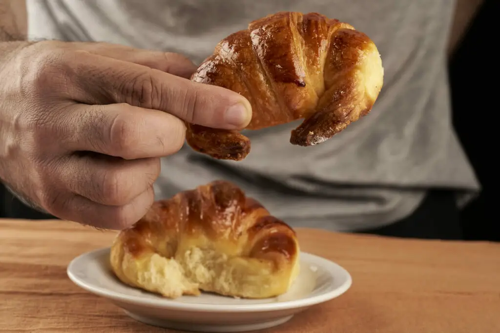 Hand holding a medialuna, a popular croissant-like food in Argentina. A person's hand is lifting a shiny, golden-brown medialuna above a plate with another medialuna.