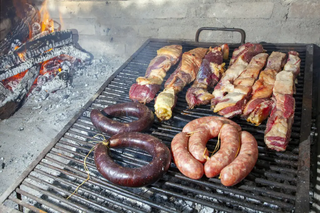 Assorted meats on a barbecue grill. Blood sausages, spiral sausages, and cuts of beef are cooking over charcoals, with visible flames from a wood fire to the left.