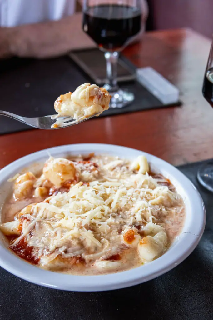 Gnocchi with sauce and cheese. A bowl of gnocchi in a creamy sauce topped with grated cheese is on a table, with a spoon lifting some gnocchi. A glass of red wine is in the background.