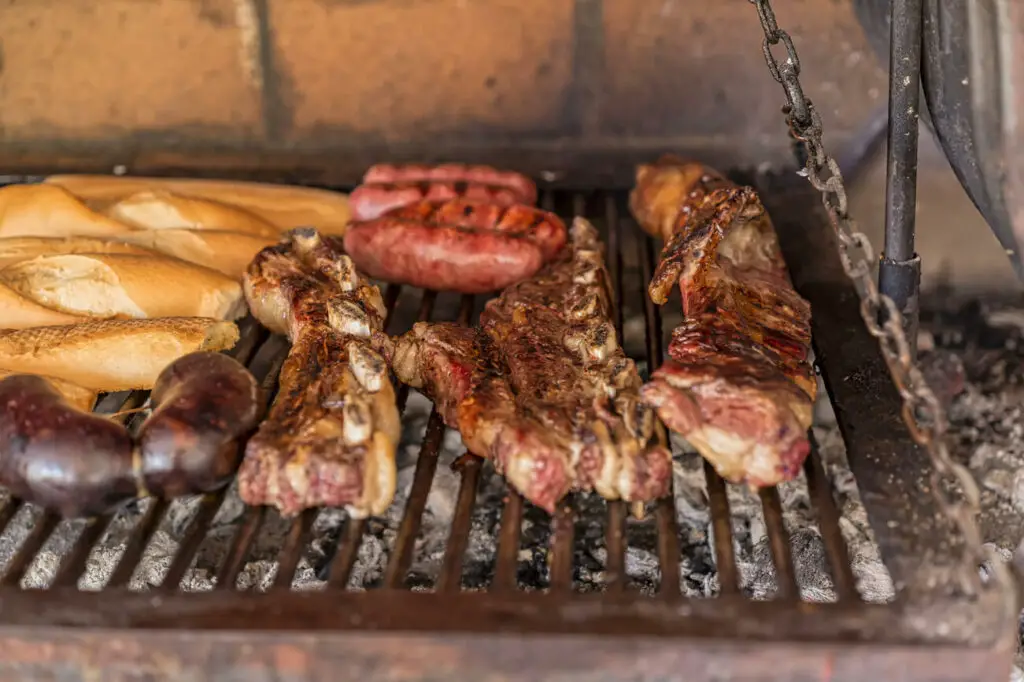 Grilled meats with bread on a barbecue. Different meats including sausages and ribs are grilling, accompanied by a row of baguettes toasting above the embers.