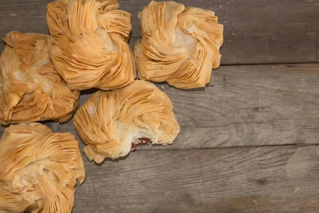 Piled flaky pastries called pastelitos on a wooden surface. Multiple square-shaped, flaky pastries are stacked, showcasing their textured layers, with some crumbs scattered around. Pastelitos are a typical Argentinian sweet.