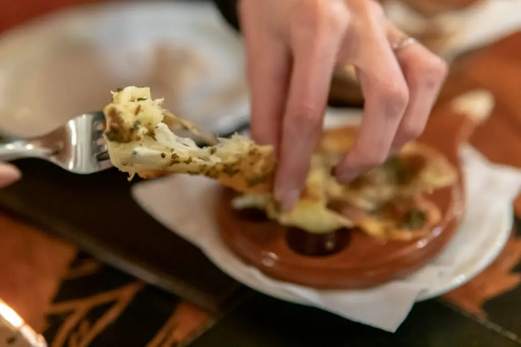 Person serving themselves food. A hand is shown grabbing provoleta cheese in Argentina with a fork from a terracotta dish, with a blurred background suggesting a dining environment.