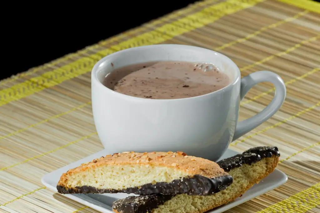 Hot chocolate with biscotti. A white cup of hot chocolate is next to almond biscotti partially dipped in dark chocolate, placed on a white plate on a bamboo mat.