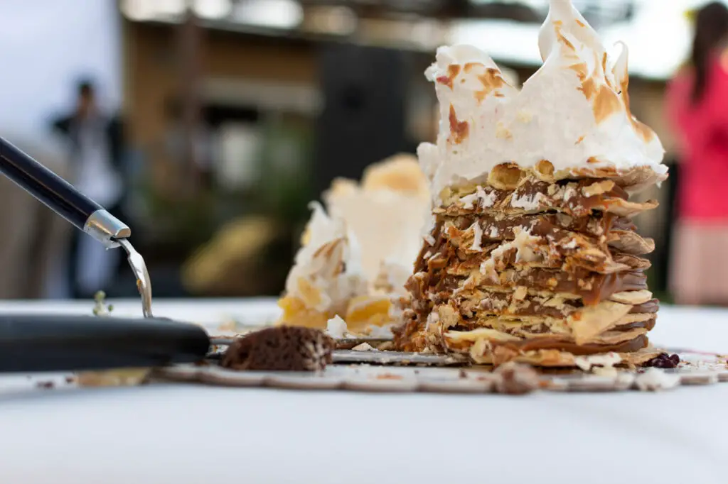 Layered pastry dessert being served. A large, multi-layered pastry called torta rogel - a popular dessert in Argentina - with a meringue top is being served with a spatula, with a blurred background indicating an outdoor setting.