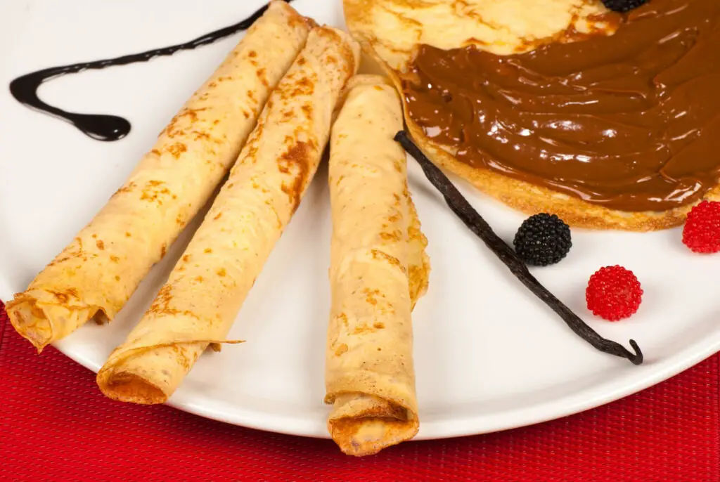 Crepes with dulce de leche and berries. Three rolled crepes and a half-covered crepe with dulce de leche are on a white plate, garnished with vanilla pods and fresh red and blackberries.