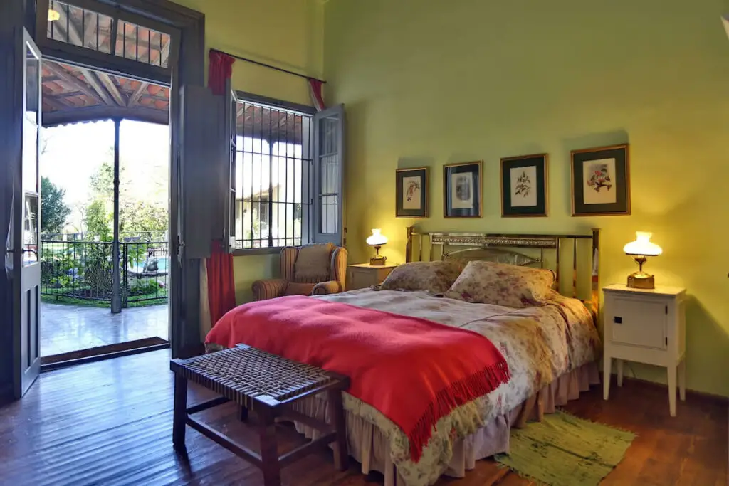 One of the cosy rooms at Estancia El Ombu near Buenos Aires. The room has a large bed covered in a red blanket, wooden floor, green walls, and soft natural lighting, giving a warm, inviting atmosphere.