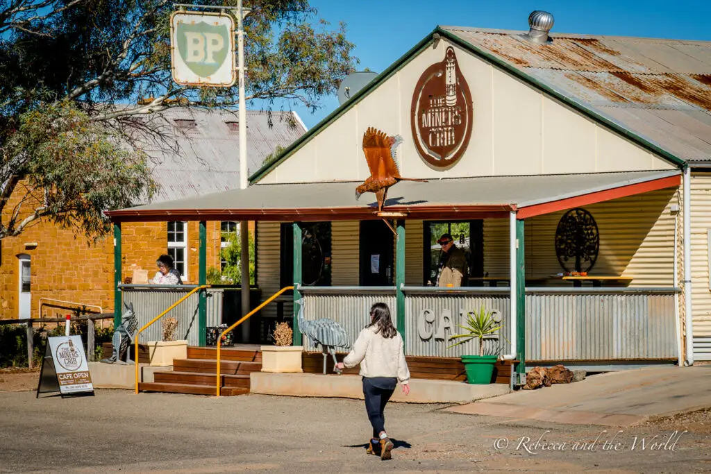 A rustic café with outdoor seating, featuring a corrugated iron roof and a vintage BP sign. A person is walking towards the entrance, and a metal bird sculpture is visible. This is the Miners Crib Cafe in Blinman, South Australia.