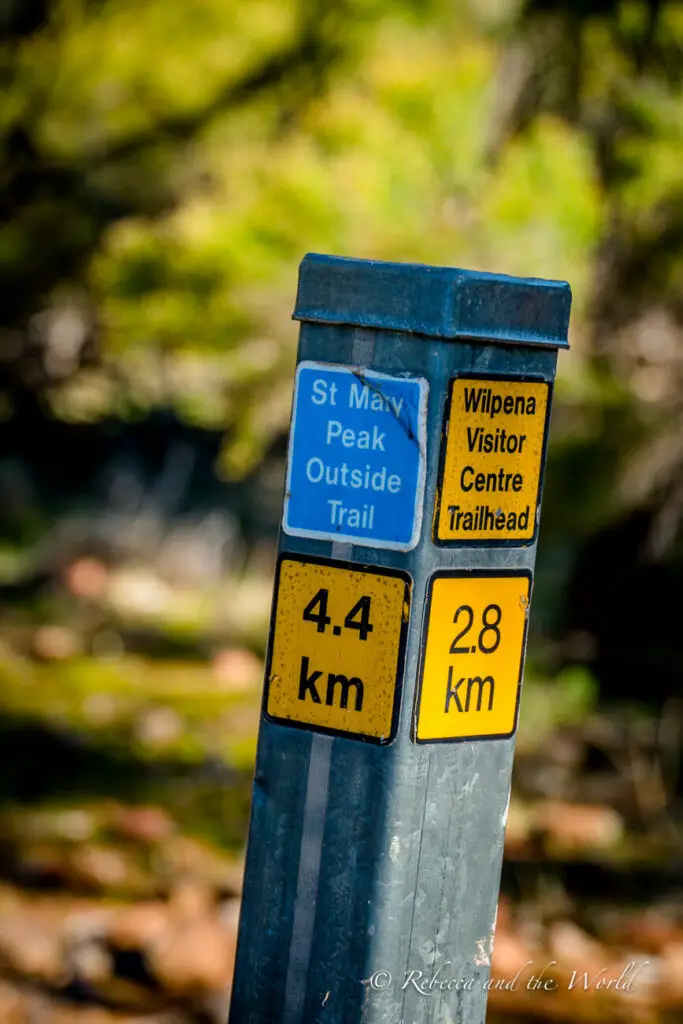 A directional trail sign with distances marked for "St Mary Peak Outside Trail" at 4.4 km and "Wilpena Visitor Centre Trailhead" at 2.8 km. The sign is blue and yellow with a blurred green background.