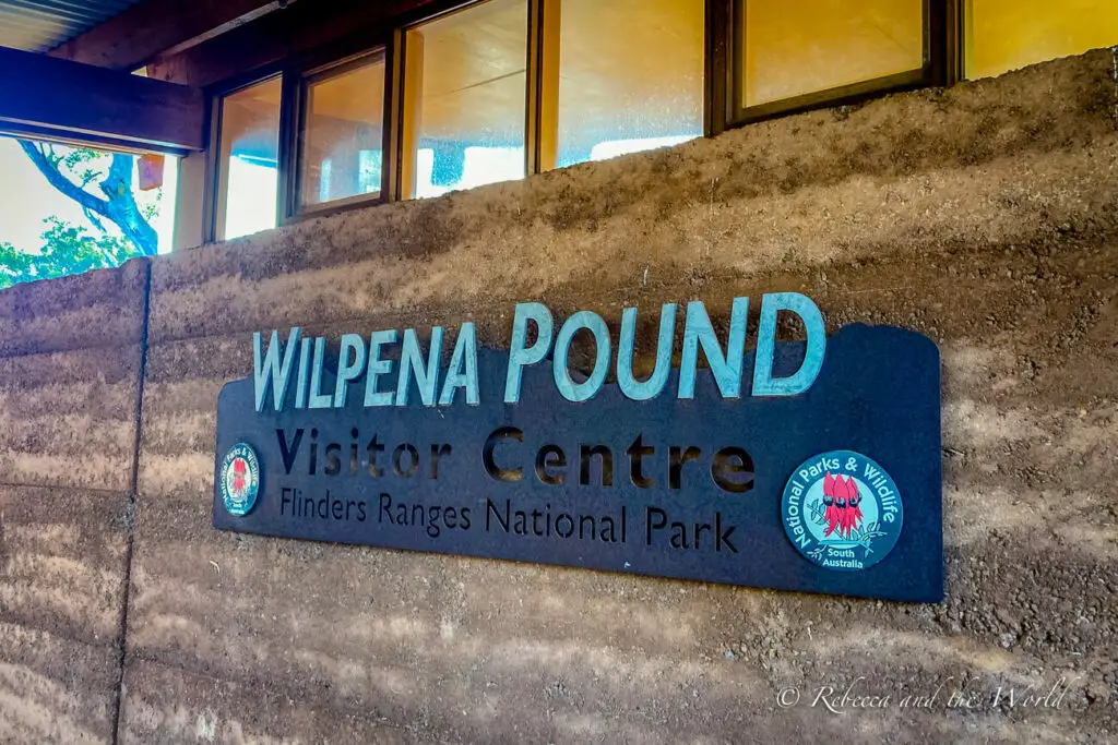 A sign reading "WILPENA POUND Visitor Centre - Flinders Ranges National Park" with the logo of South Australia National Parks & Wildlife. The sign is mounted on a concrete wall with a window in the background. The Ikara Flinders Ranges National Park Visitor Information Centre sign - the centre is located within Wilpena Pound Resort and it's one of the first places you should visit when you arrive at the national park.