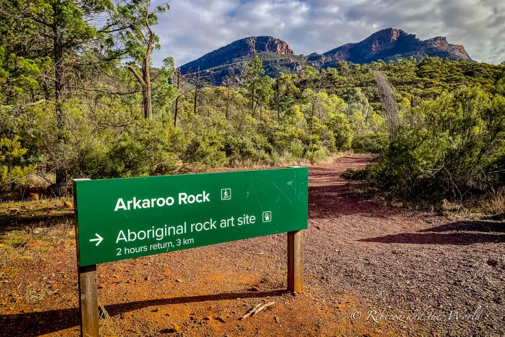 A green sign indicating "Arkaroо Rock - Aboriginal rock art site" with an arrow pointing to the right. It mentions a 2-hour return and 3 km distance. The background shows a dirt path leading towards wooded hills. This is a great hike in Flinders Ranges National Park.