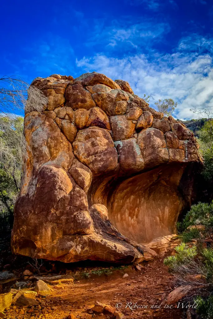 A large, freestanding rock formation with a hollowed-out area resembling a cave. The rock has a rounded appearance with reddish-brown tones, set against a blue sky with light clouds. This is one of the rocks you'll see on the Arkaroo Rock hike at Flinders Ranges National Park.