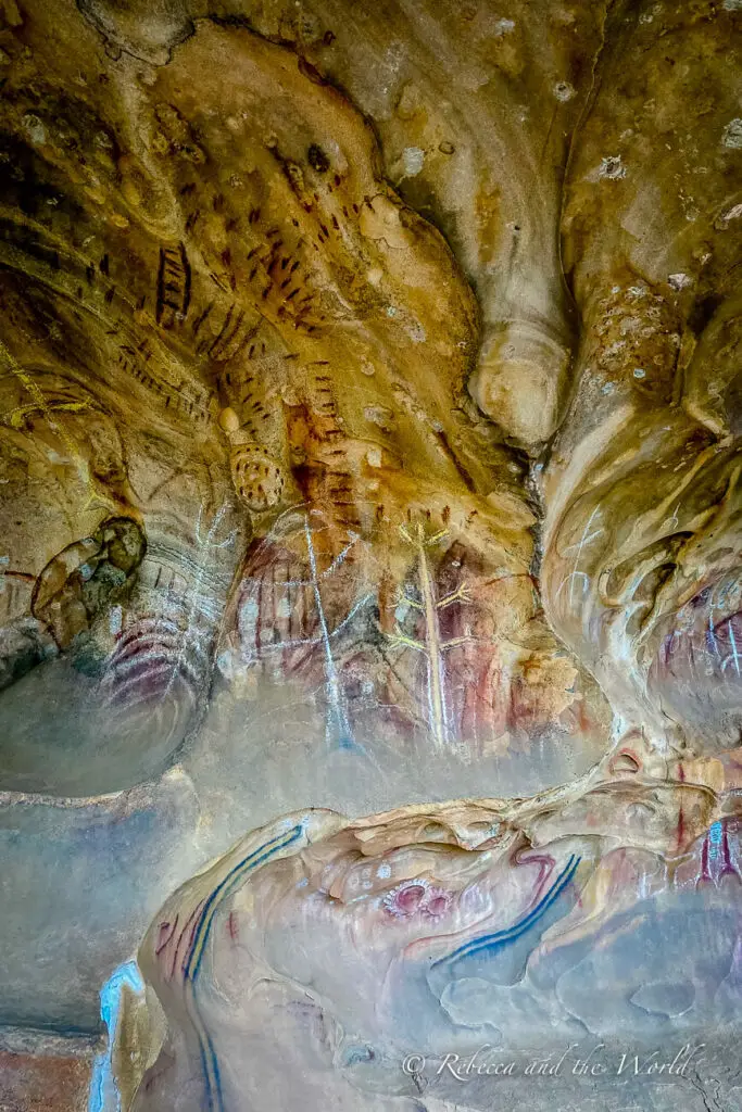A close-up of ancient rock art seen on a cave wall in Flinders Ranges National Park, featuring patterns and animal figures in red, white, and yellow ochre. The natural contours of the rock surface enhance the depth of the paintings.