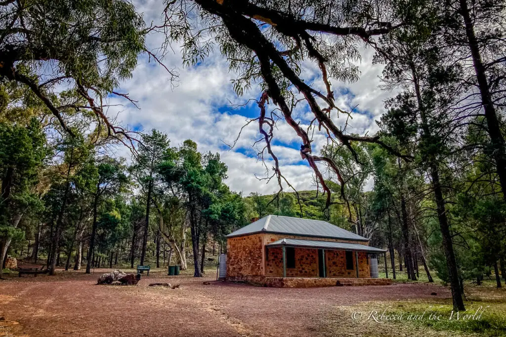 A small, rustic stone building with a metal roof, situated in a clearing surrounded by dense woodland. The sky is partially cloudy and the ground is covered with red earth. This is the homestead you'll see on the Hills Homestead hike near Wilpena Pound in the Flinders Ranges National Park.